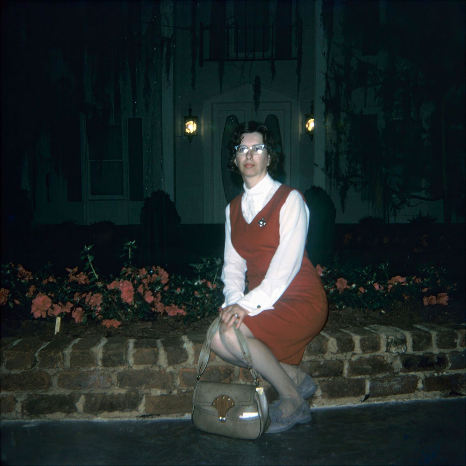 Woman in red vest and white blouse sitting on brick wall at night with flowers and lit building behind her.