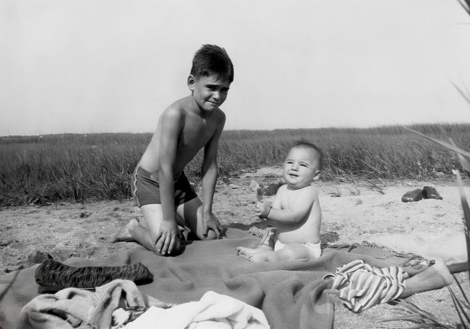 Black and white photo of a smiling boy and baby sitting on towels at a sandy beach near tall grass.