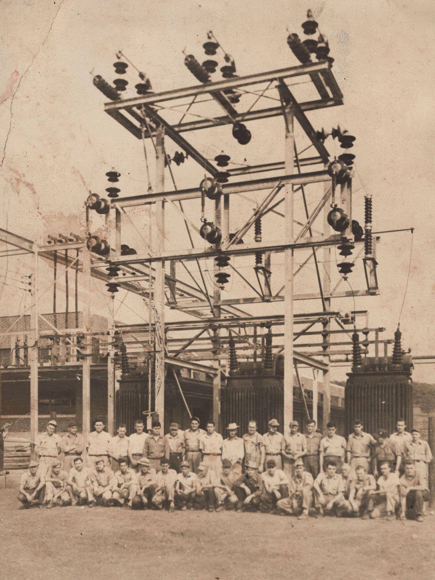 Vintage sepia photo of workers posing before an electrical substation with high-voltage insulators and steel framework.