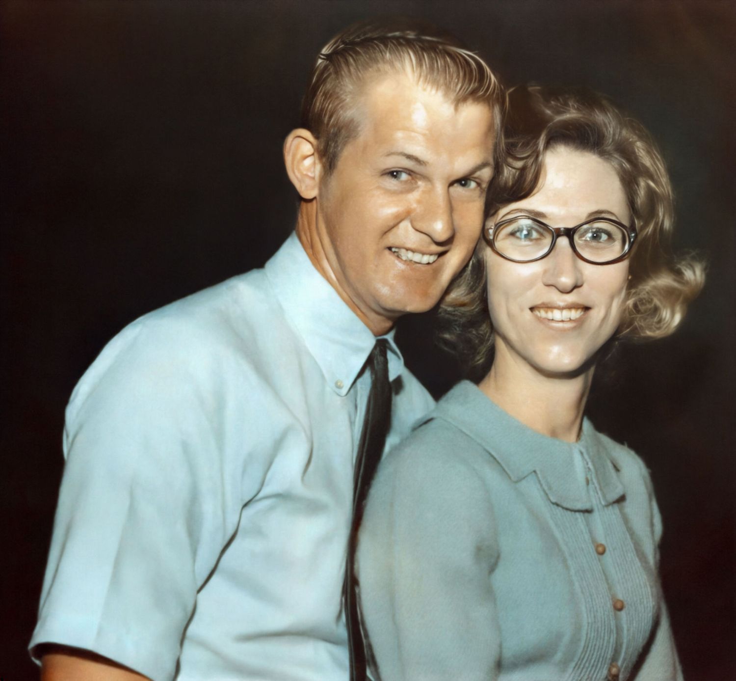 Vintage 1960s color portrait of a smiling couple, man in blue shirt with tie and woman wearing cat-eye glasses.