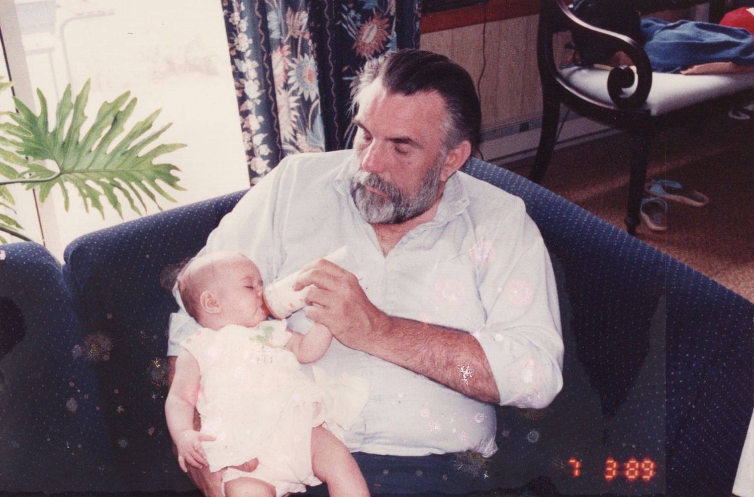 Elderly bearded man bottle-feeding a newborn baby while seated in a blue armchair, vintage 1989 photo.