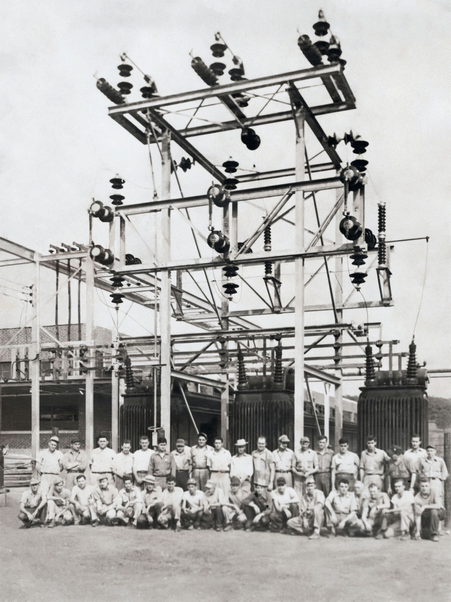 Vintage black and white photo of workers posing at an electrical substation with high-voltage equipment and insulators.