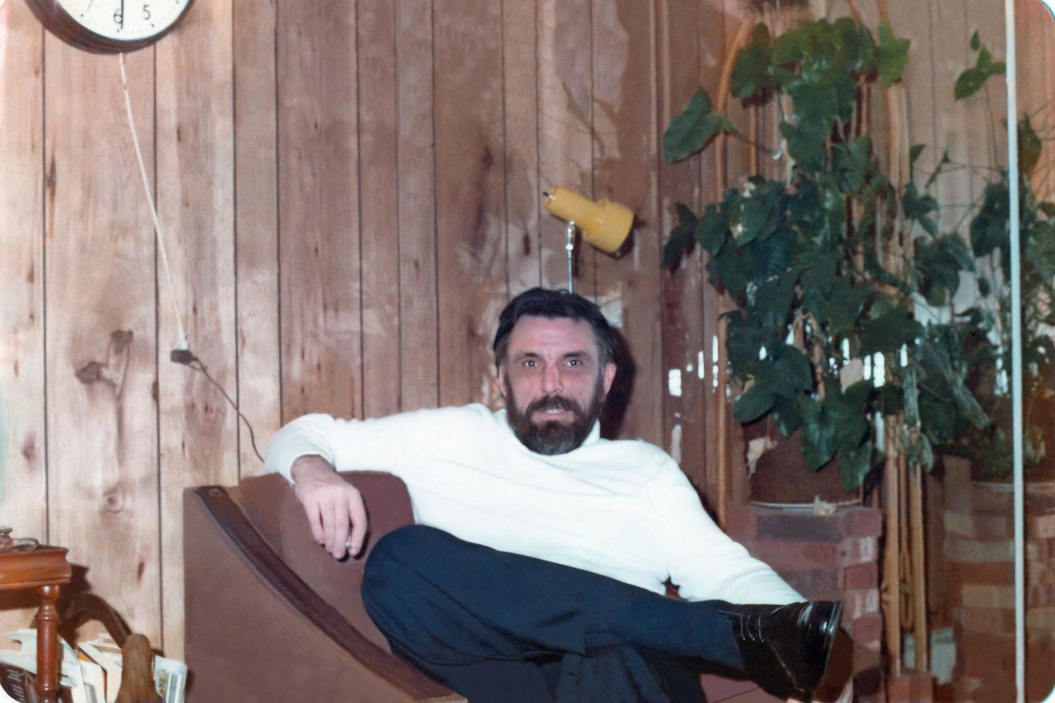 Bearded man relaxing in a chair inside a wood-paneled room with plants and a clock on the wall.