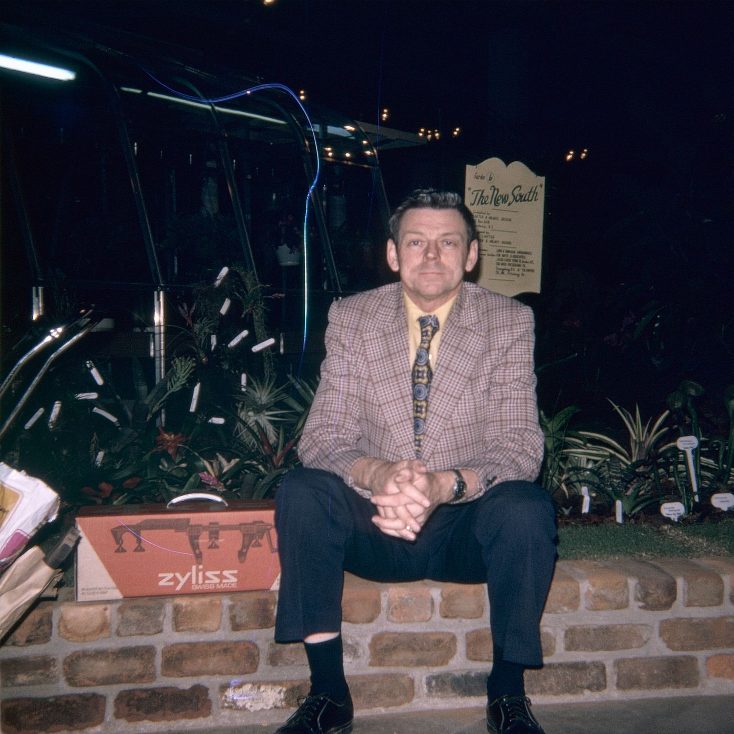 Man in plaid blazer sitting on brick wall surrounded by tropical plants near a Zyliss display at an outdoor event.