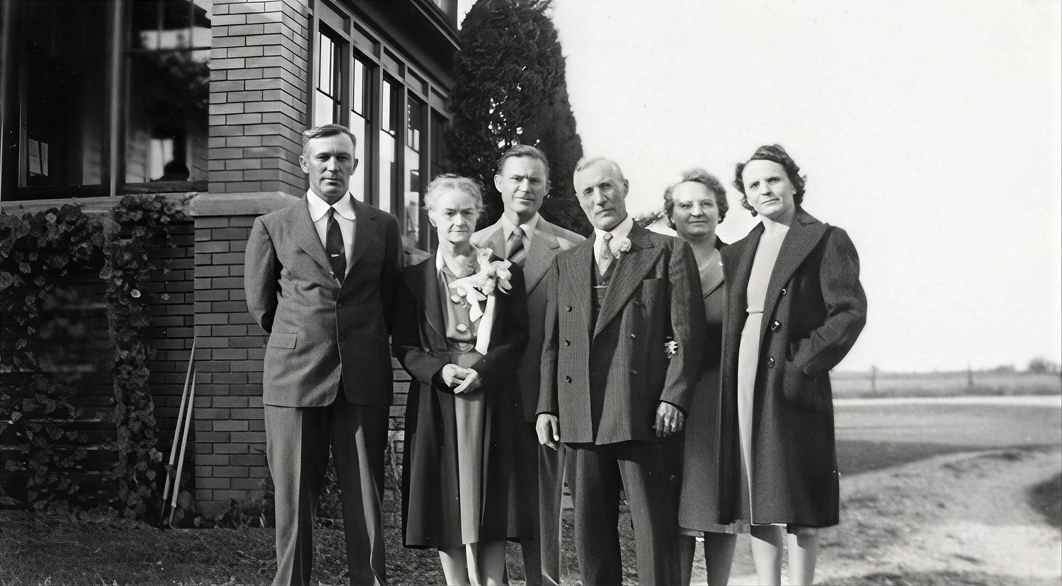 Vintage black and white photo of a family group of seven adults posing outdoors near a brick building.