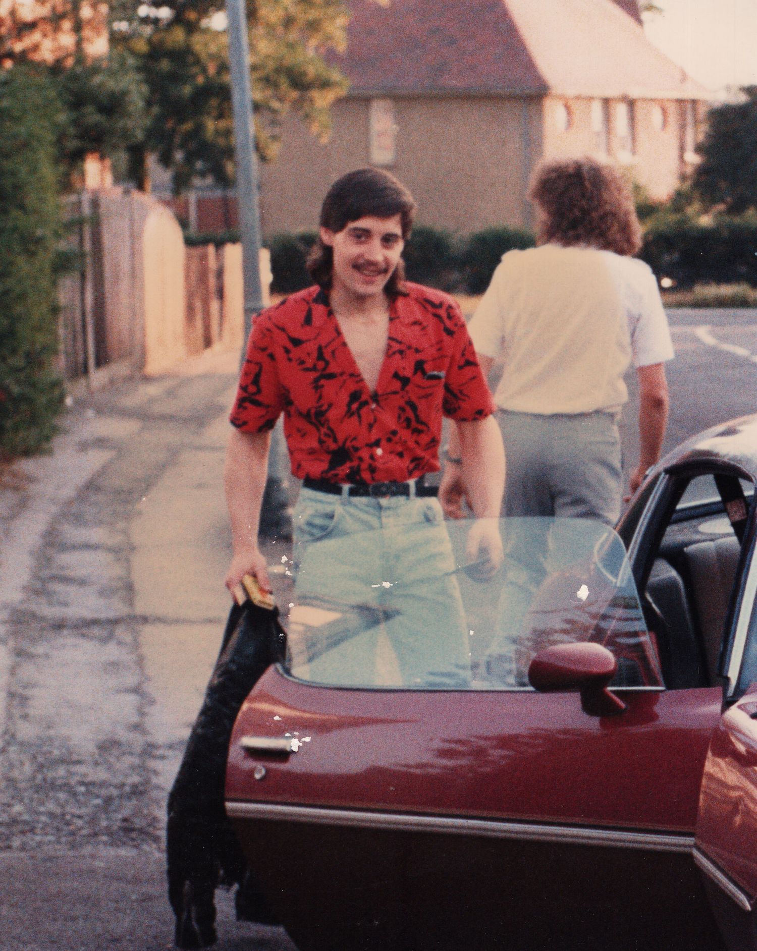 A man in a red patterned shirt and jeans leans on a vintage car on a residential street in the 1980s.