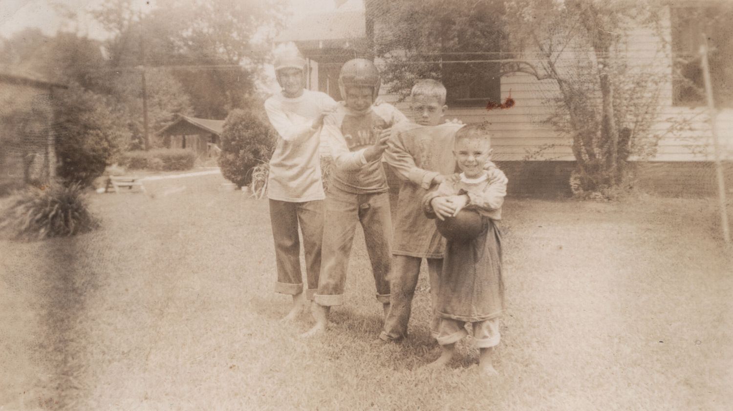 Vintage sepia photo of four children posing together outdoors near a house, likely from the mid-20th century.