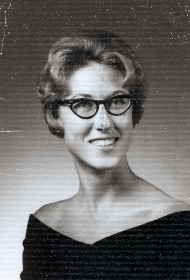Vintage black and white portrait of a young woman with cat-eye glasses and bouffant hairstyle, circa 1960s.