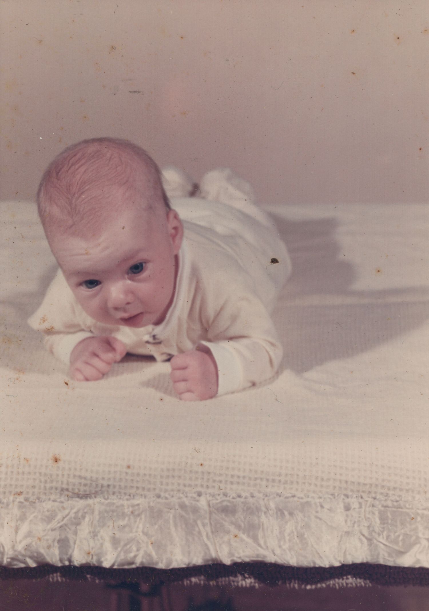 Vintage photo of a newborn baby lying on stomach on a white textured blanket, lifting head and looking at camera.