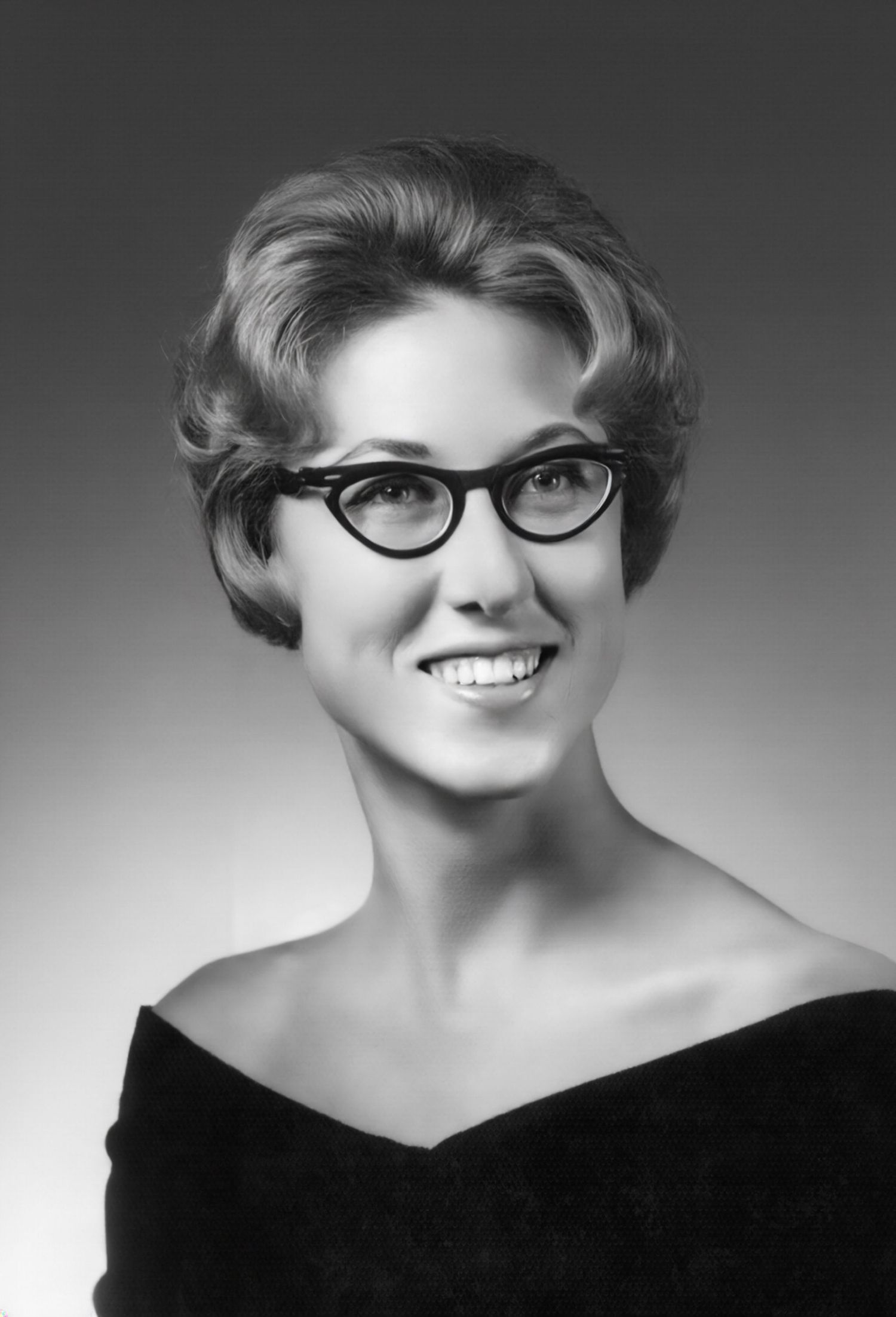 Black and white portrait of a smiling young woman with short curly hair wearing cat-eye glasses and an off-shoulder top.
