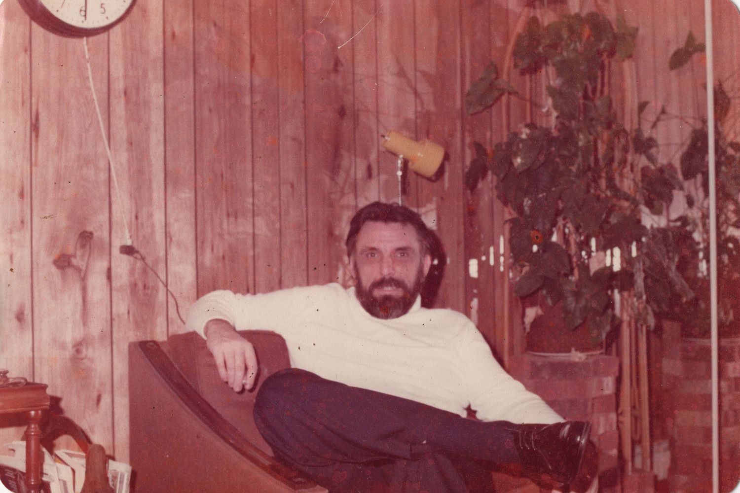Vintage 1970s photo of a bearded man relaxing in a chair beside wood-paneled walls and indoor plants.