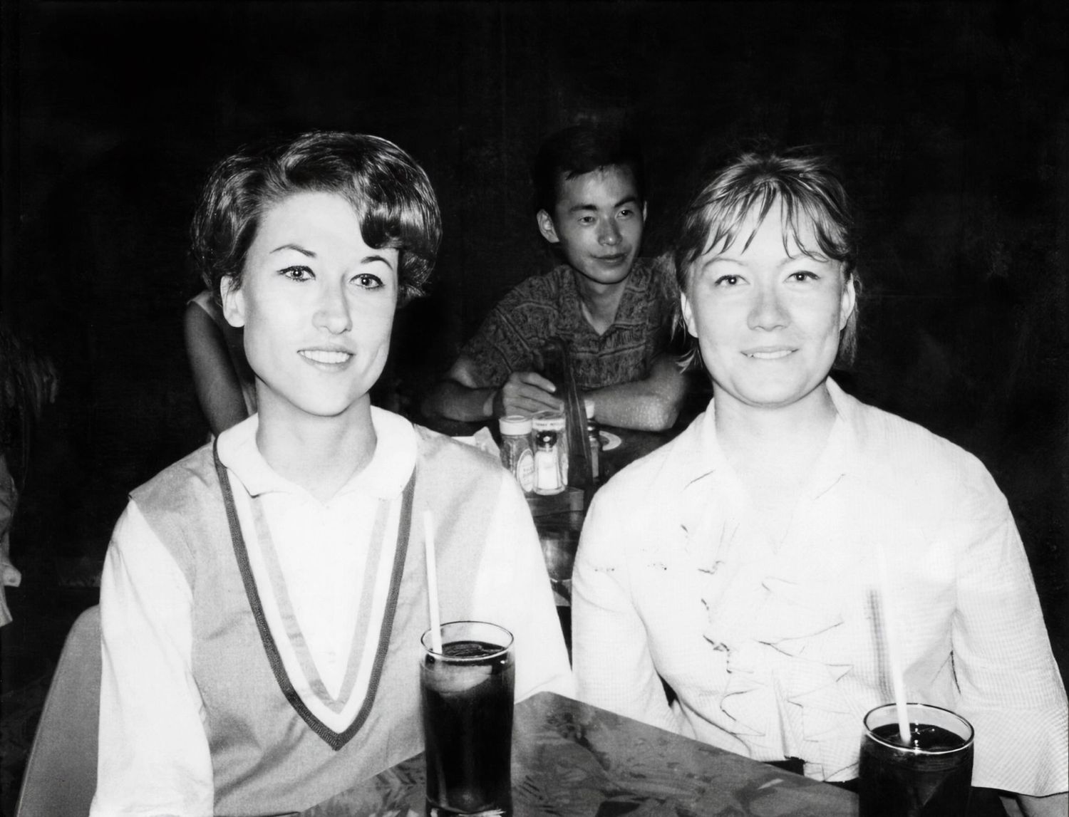 Two women sitting at a table with drinks in a vintage black and white photo, with a young boy visible in the background.