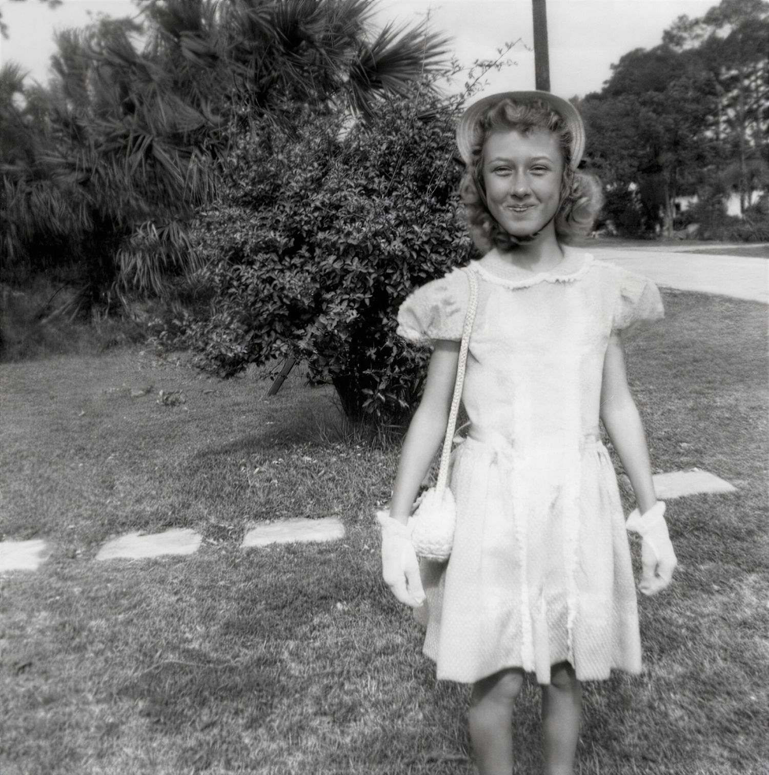 Vintage black and white photo of a young girl in a white dress and gloves standing on a lawn near trees.