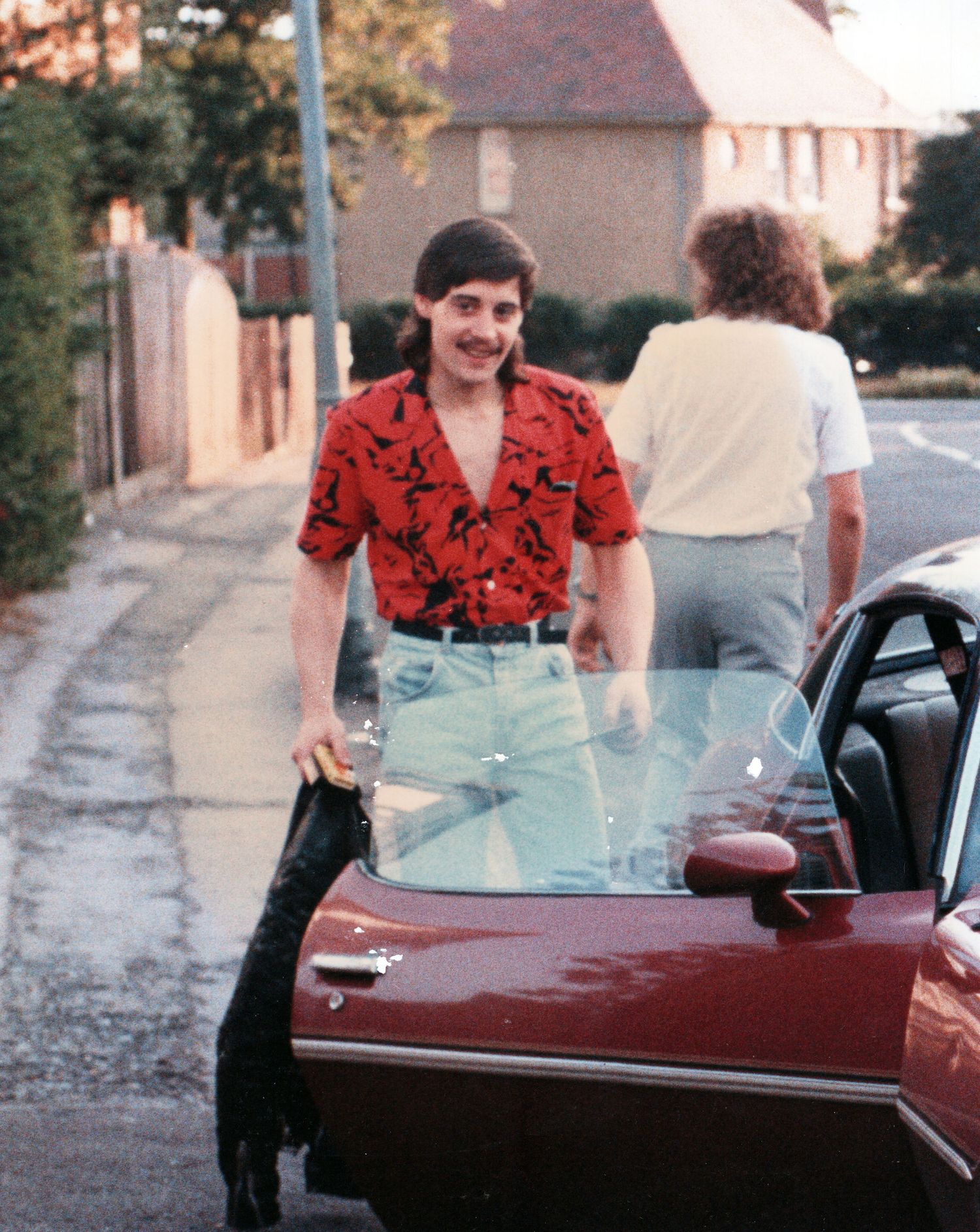 Man in red patterned shirt leaning on a vintage red car on a residential street, with another person standing nearby.