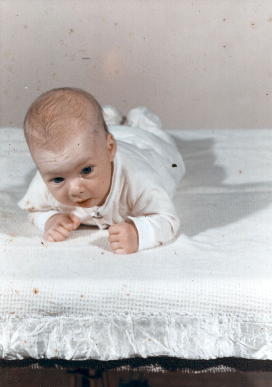 A baby lying on their tummy on a white textured blanket, lifting their head and looking at the camera.