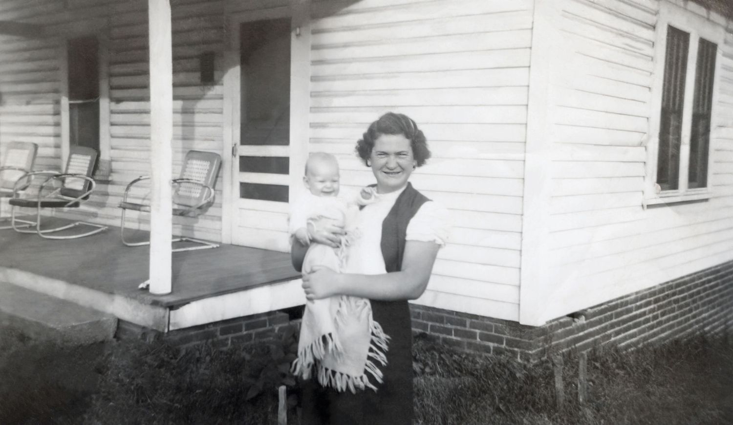 Vintage black and white photo of a smiling woman holding a baby outside a white clapboard house with porch chairs.