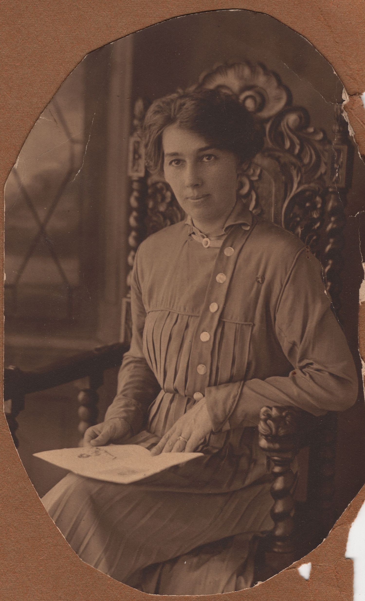 Vintage sepia portrait of a young woman seated in ornate chair, wearing button-front dress, holding paper, circa early 1900s.