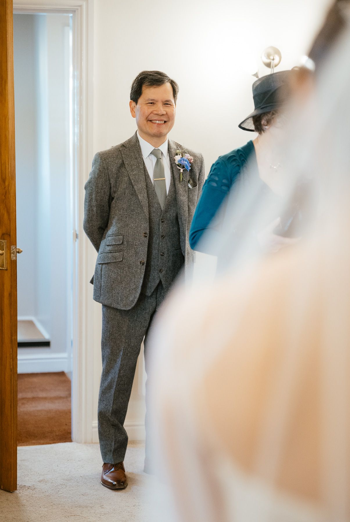 A groom wearing a classic grey three-piece suit smiles while standing in a doorway.