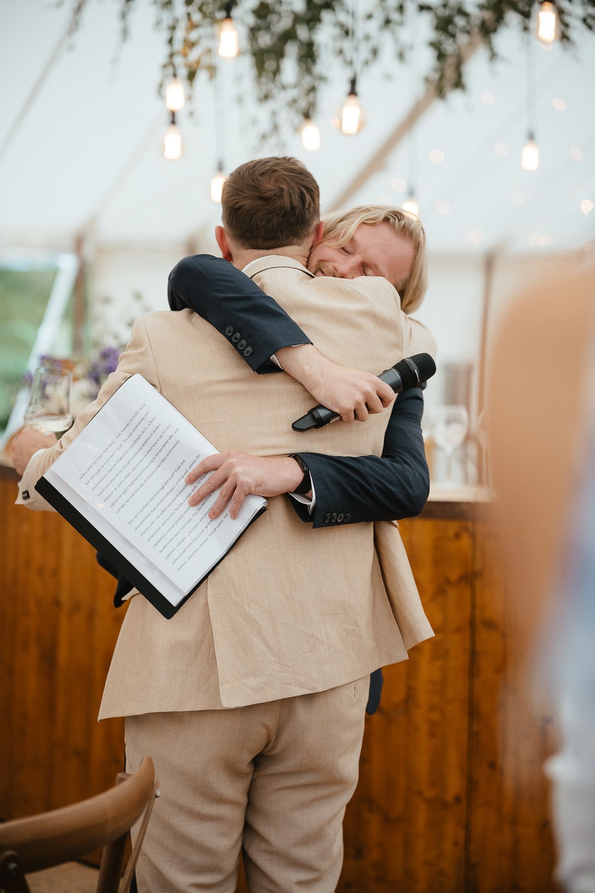 Two people share an emotional embrace at a wedding ceremony underneath strings of glowing lights in an indoor venue.