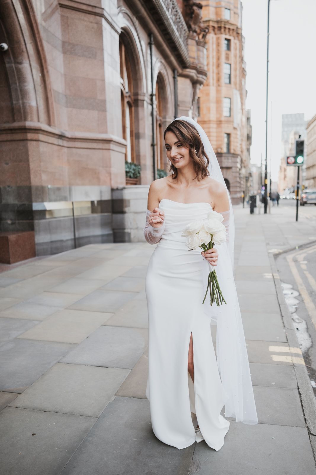 Bride in elegant off-shoulder white wedding dress holds small bouquet while standing on urban street near historic building.