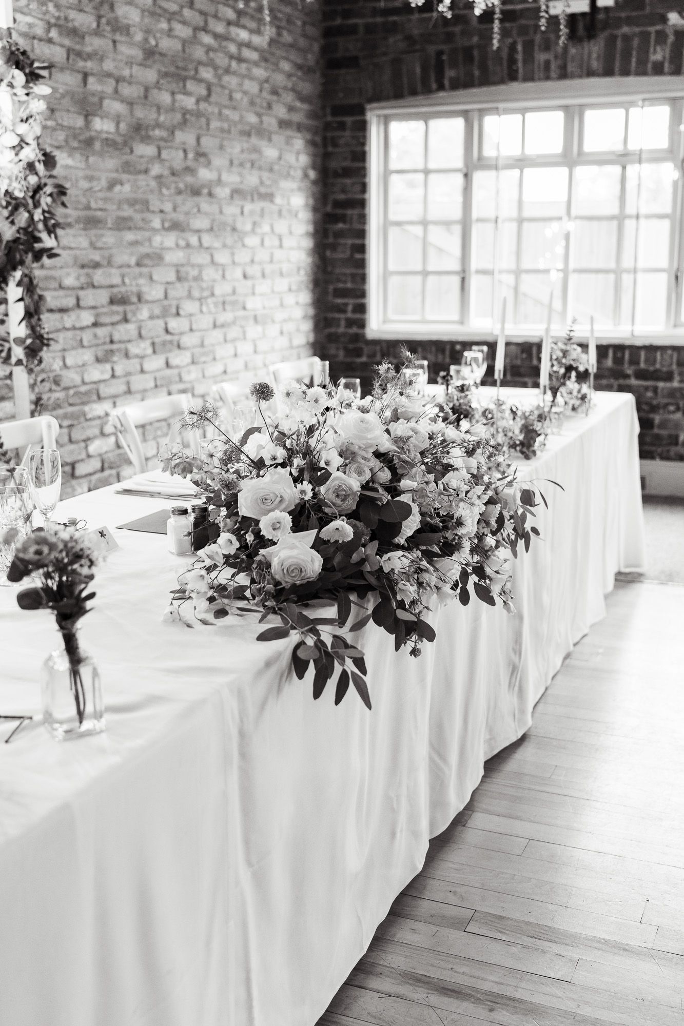 Black and white photo of an elegant wedding reception table with lush floral centerpieces and white linens in brick-walled venue.