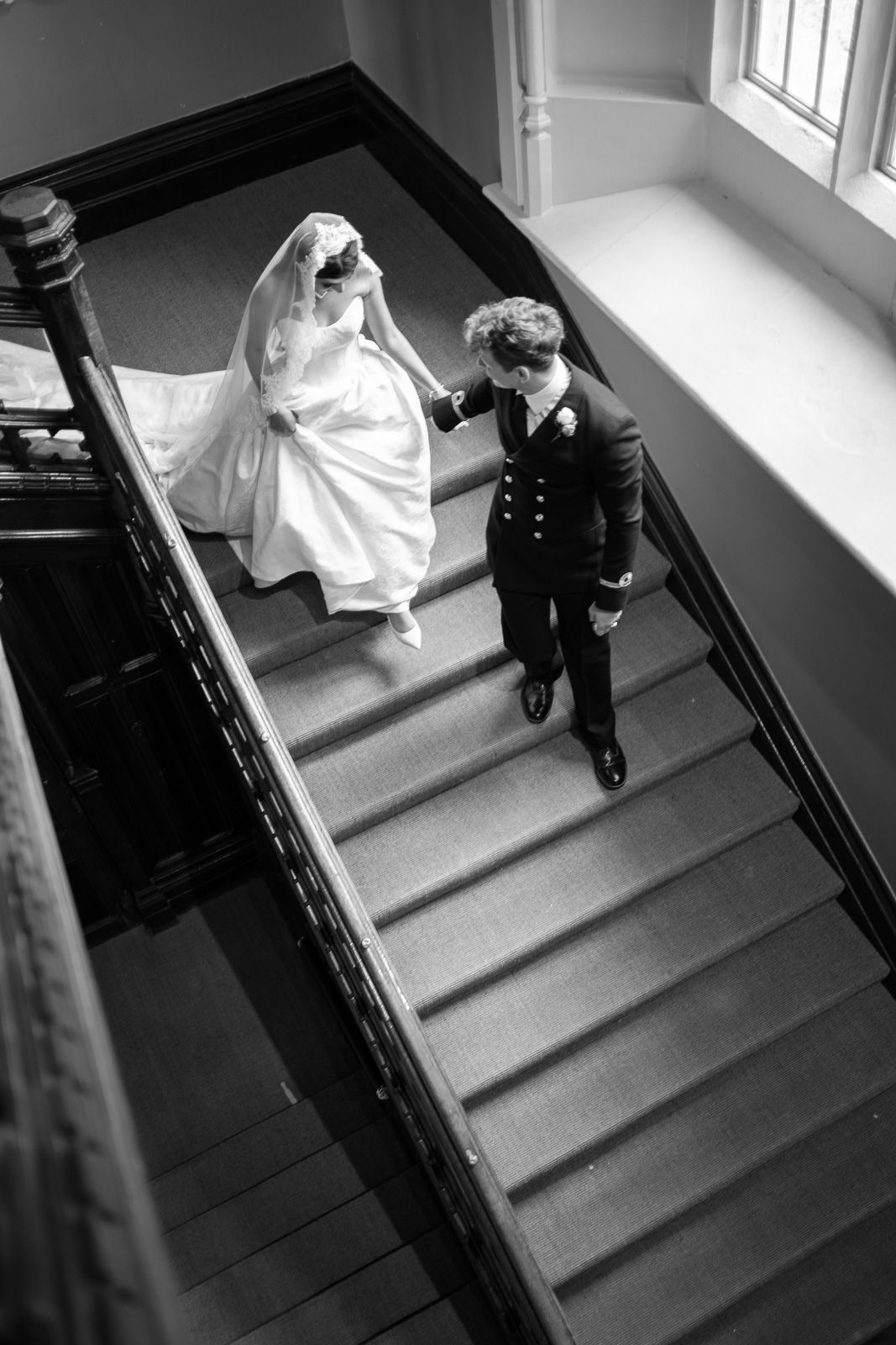 Black and white photo of bride and groom walking down ornate staircase, holding hands.