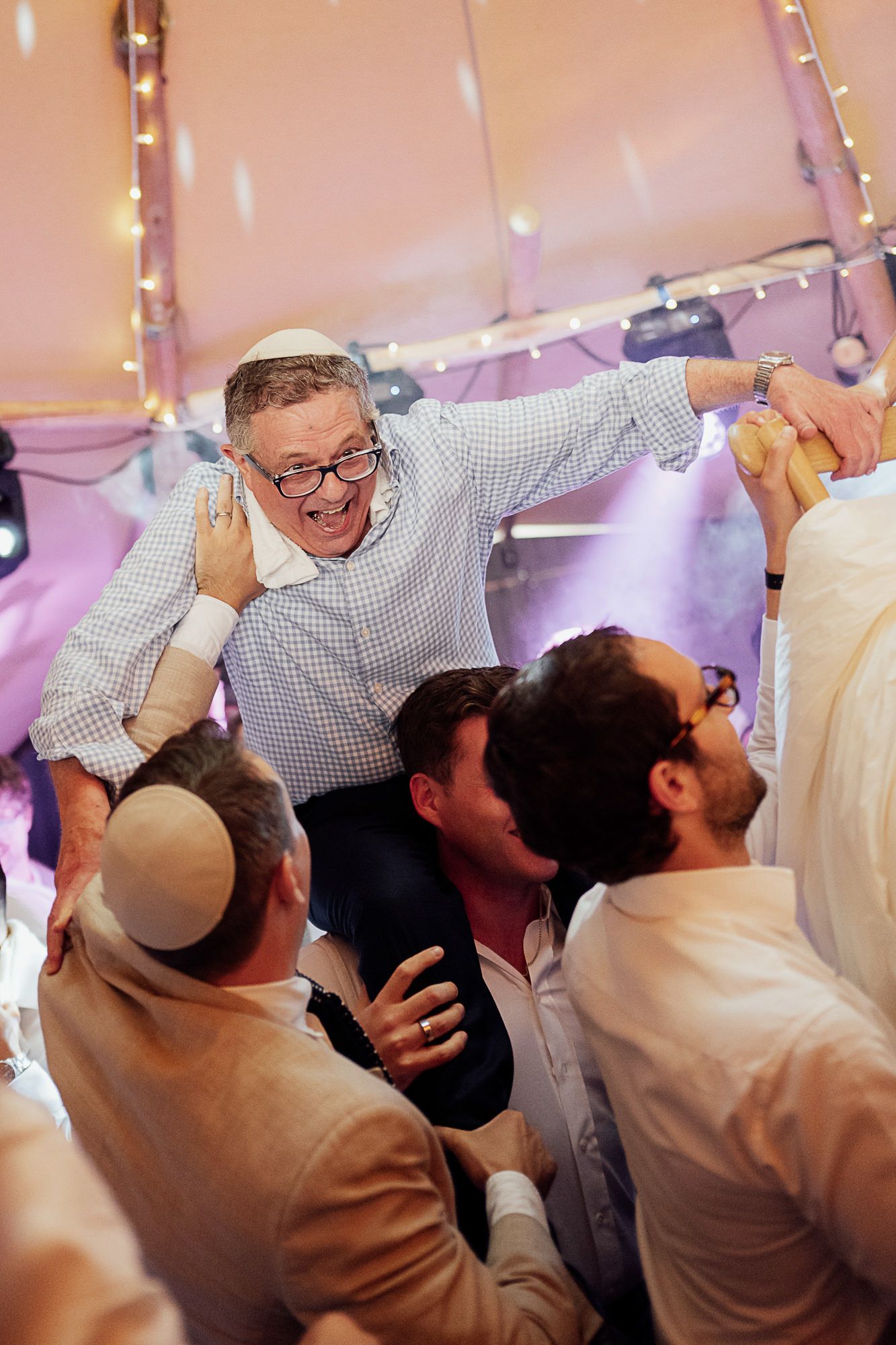 Joyful man in kippah lifted by friends during lively Jewish wedding celebration under a festive tent.