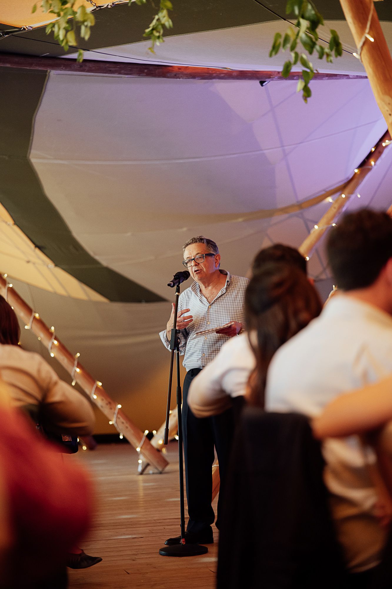 Man speaking at microphone stand inside decorated tent venue with fairy lights and draped fabric ceiling.