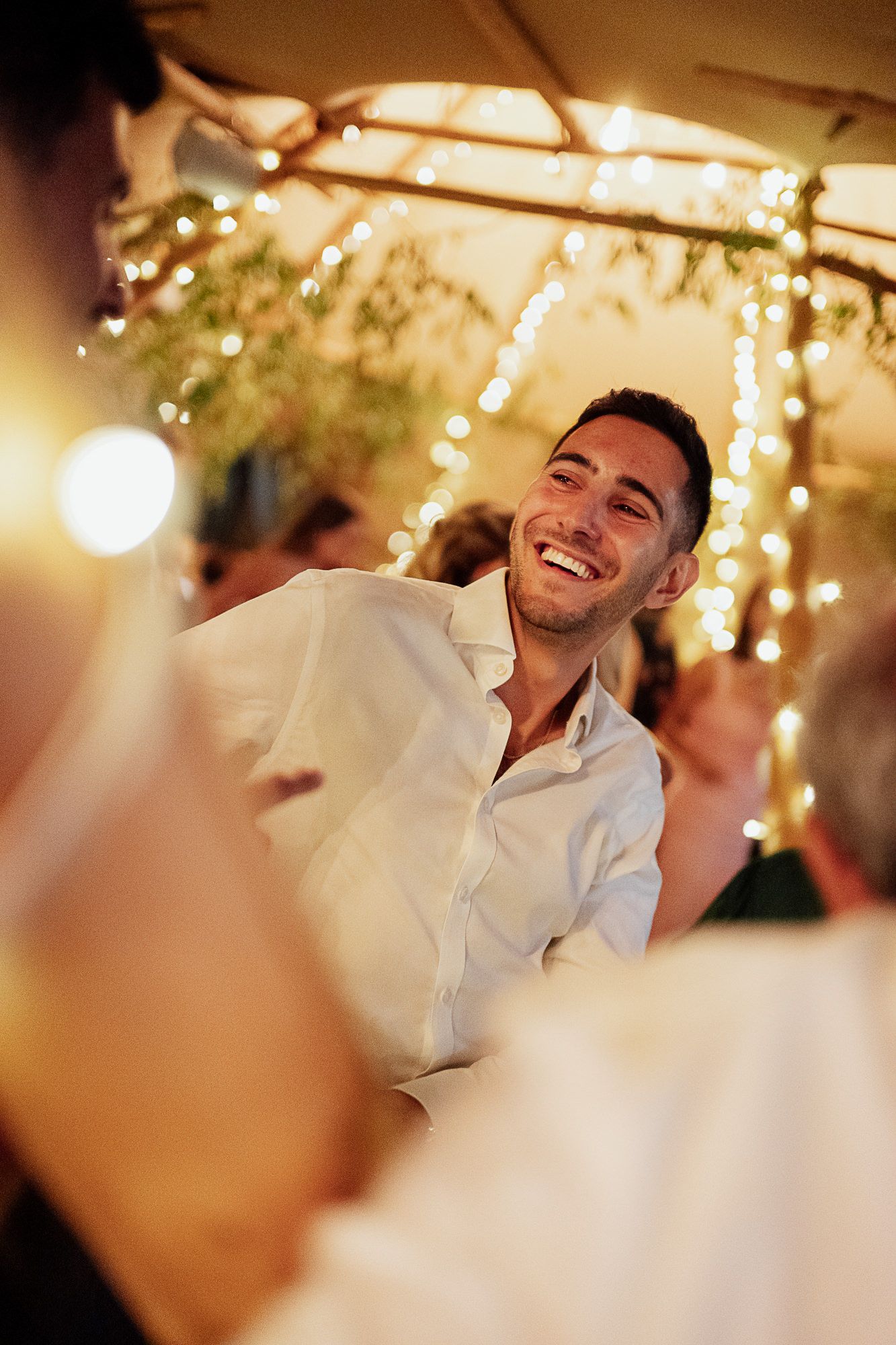 Smiling young man in white shirt laughing at outdoor evening party with fairy lights in background.