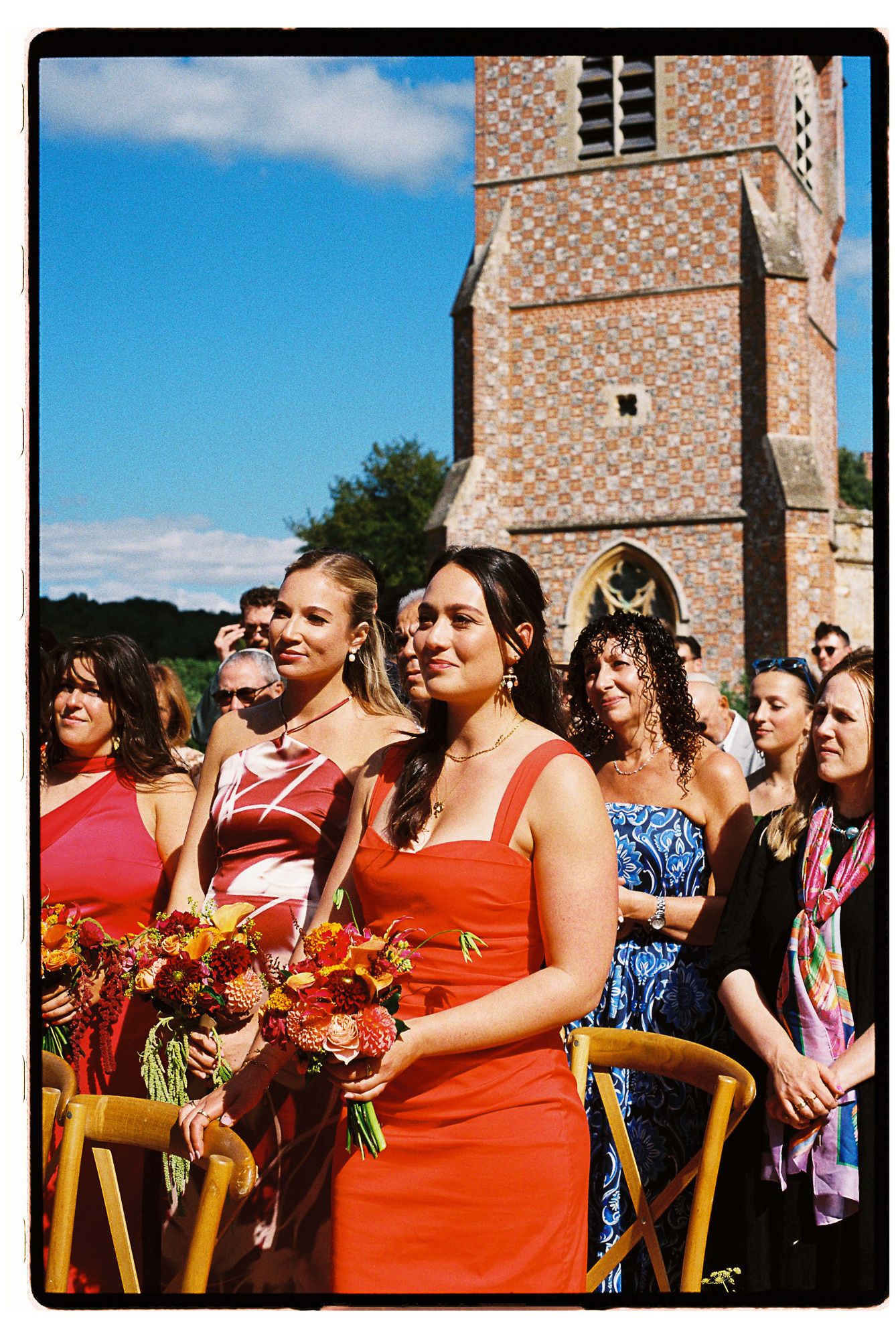 Bridesmaids in red dresses holding floral bouquets stand outdoors at a wedding ceremony near a historic brick church.