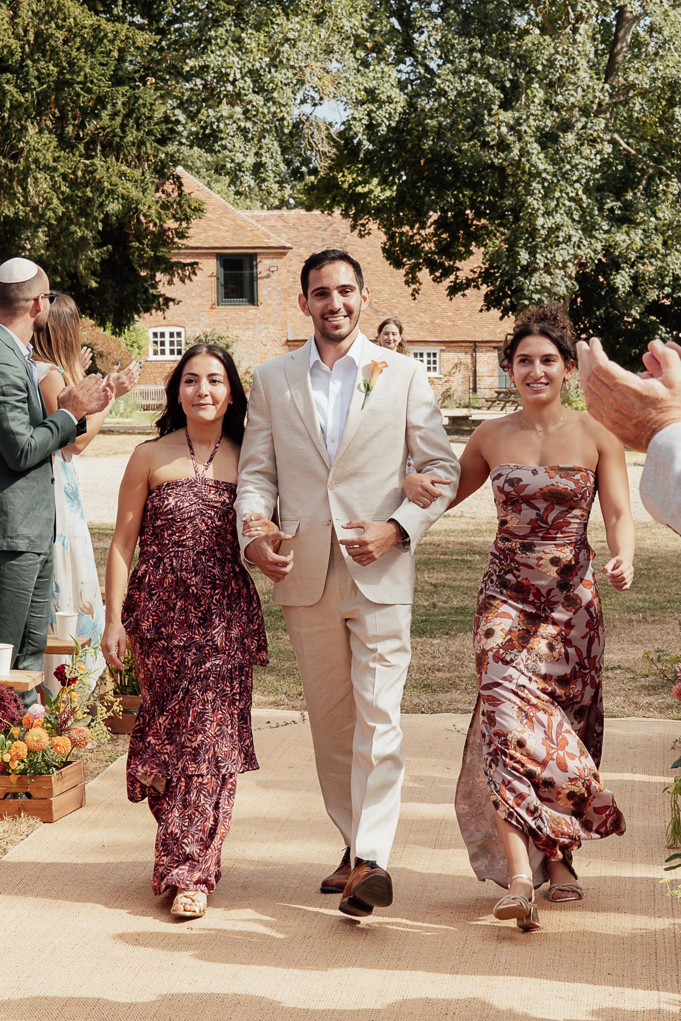 Groom in beige suit walks with two women in floral dresses at an outdoor wedding ceremony.