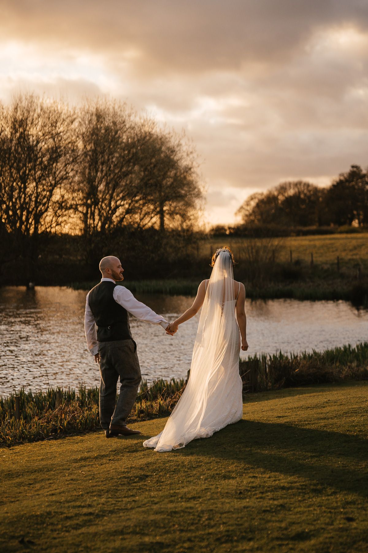Two newlyweds hold hands by a lake at golden hour as sunlight filters through clouds creating a romantic atmosphere.