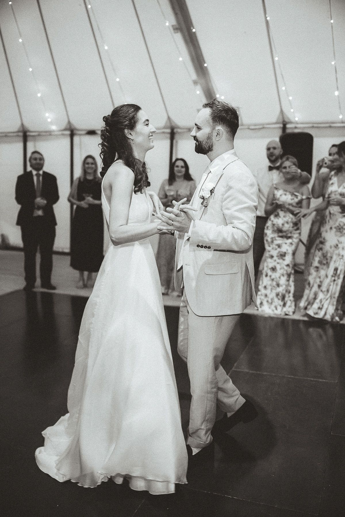 Black and white photo of a newlywed couple sharing their first dance at a wedding reception under a tent with string lights.