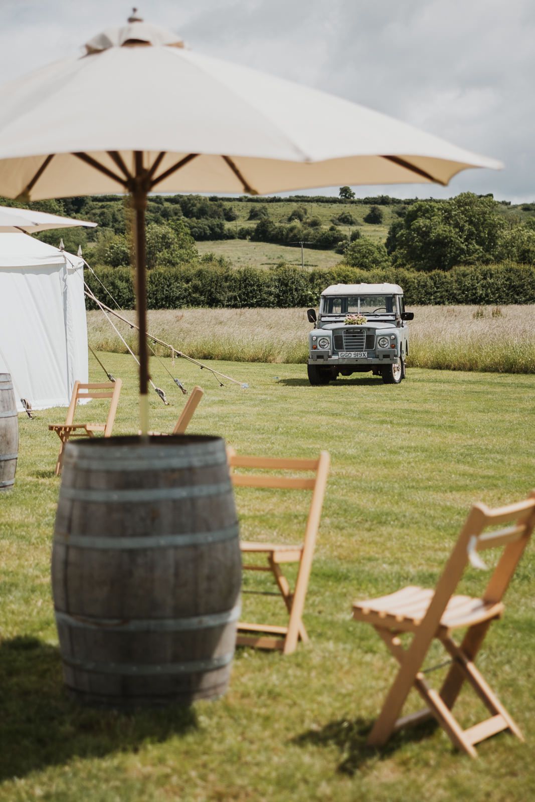 Vintage Land Rover parked on green lawn with wooden barrel, folding chairs and beige umbrella at outdoor event setup.