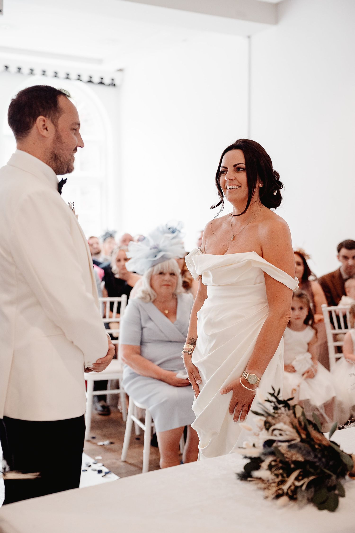 A bride and groom share their first dance while wedding guests look on in a bright white room.