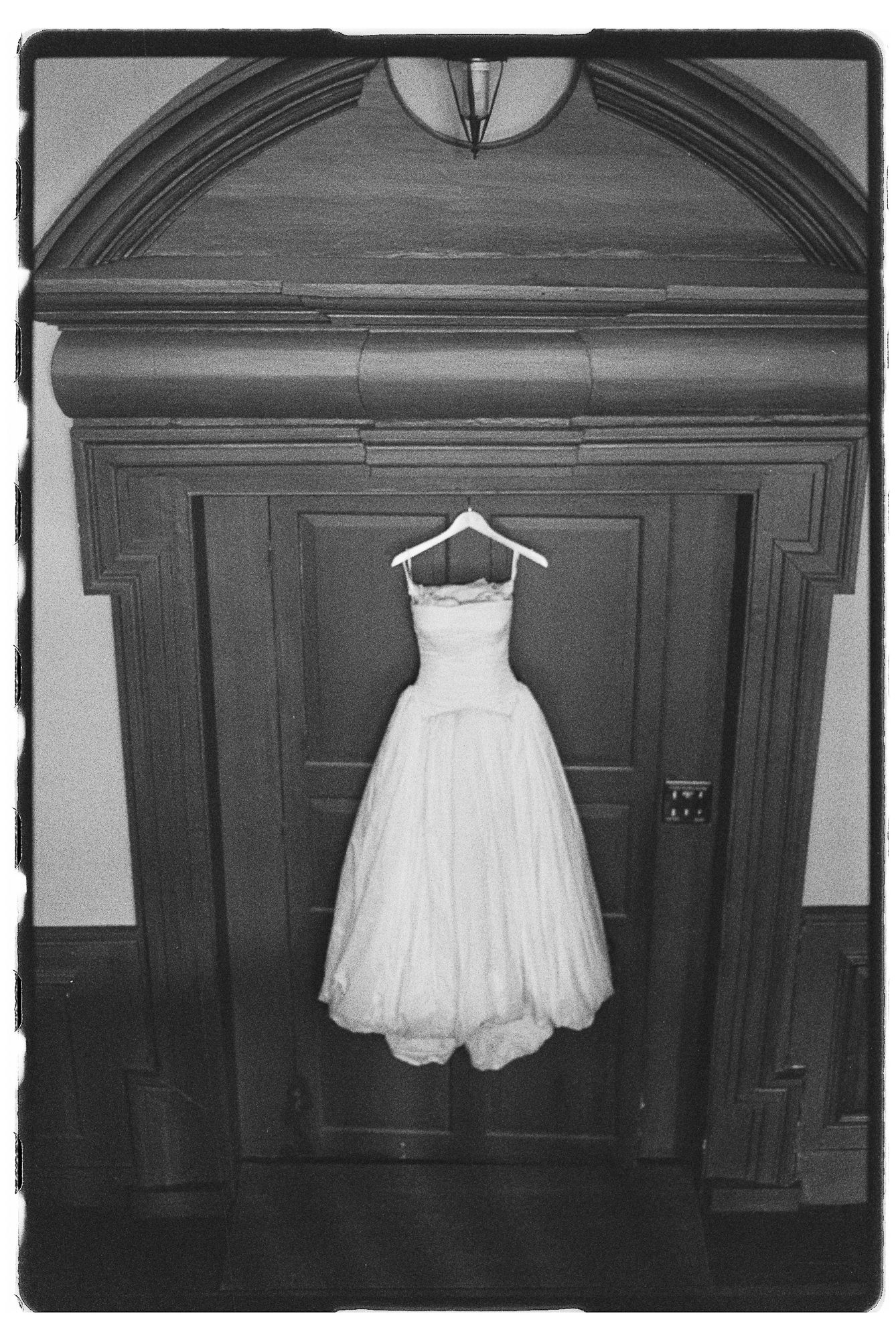 Black and white photo of an elegant wedding dress hanging on a hanger against a grand wooden door.