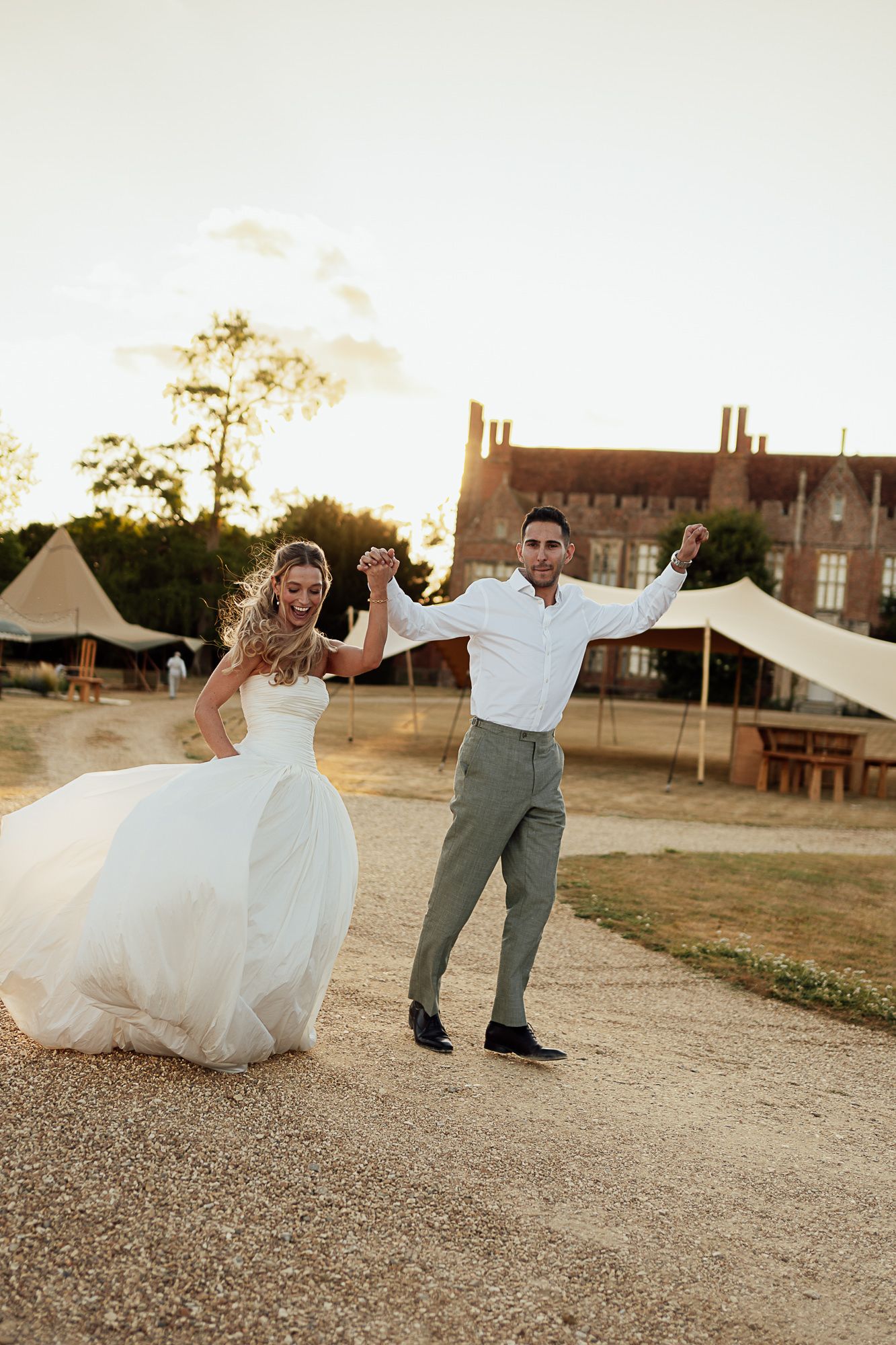 Bride and groom dancing joyfully outdoors at golden hour with a historic manor and wedding tents in the background.