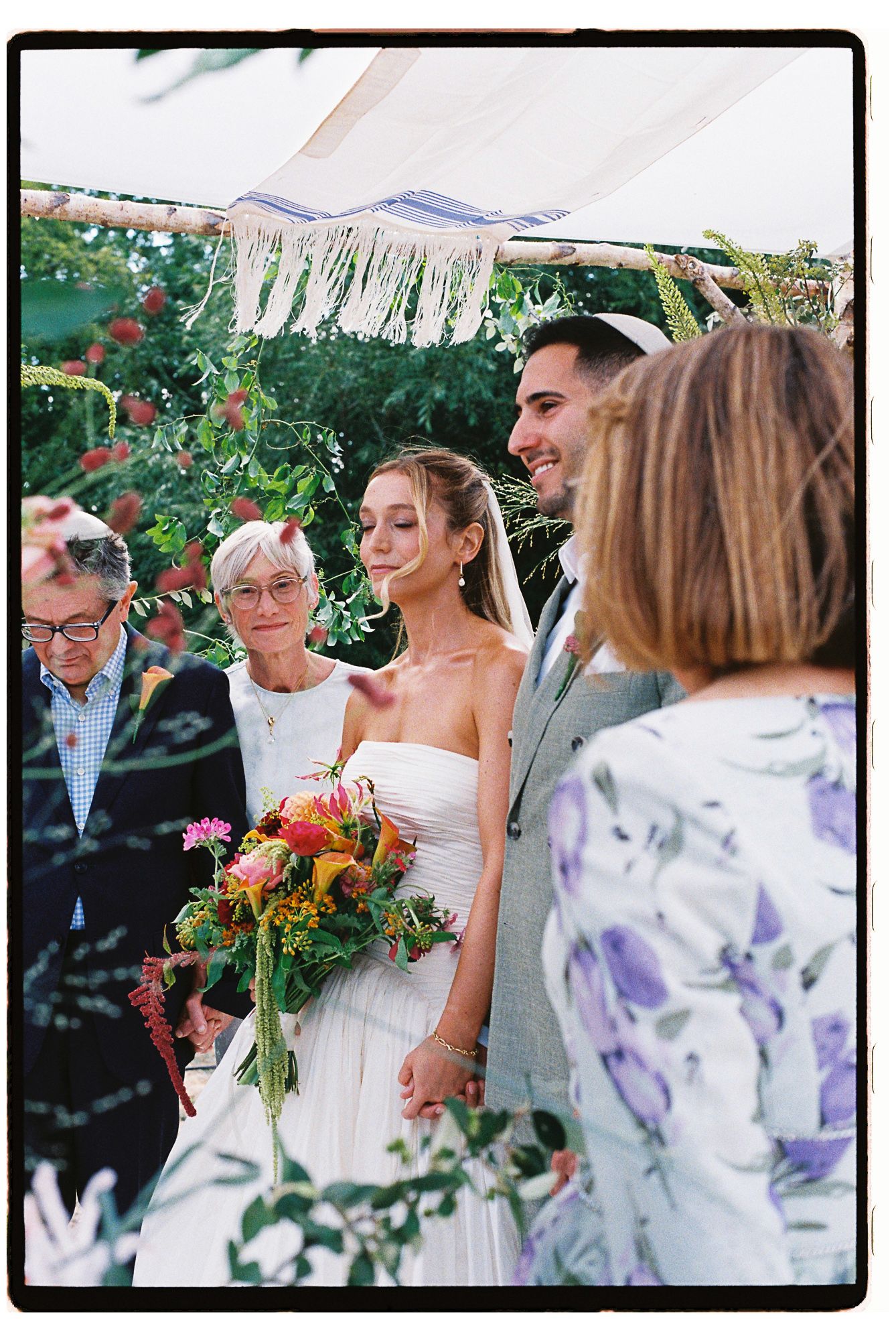 Bride holding colorful bouquet stands with groom under floral chuppah during outdoor Jewish wedding ceremony.
