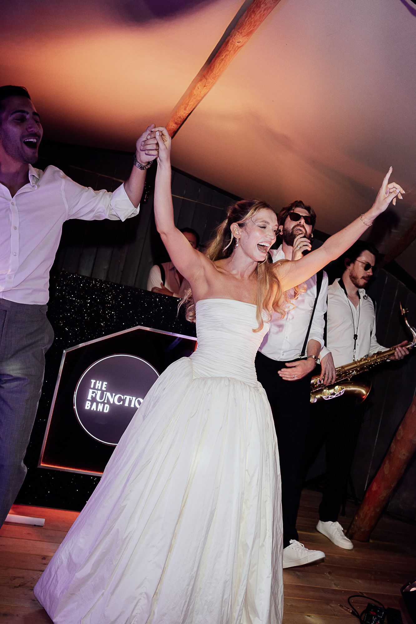 Bride in white gown dancing joyfully with guests and live function band performing on stage at wedding reception.