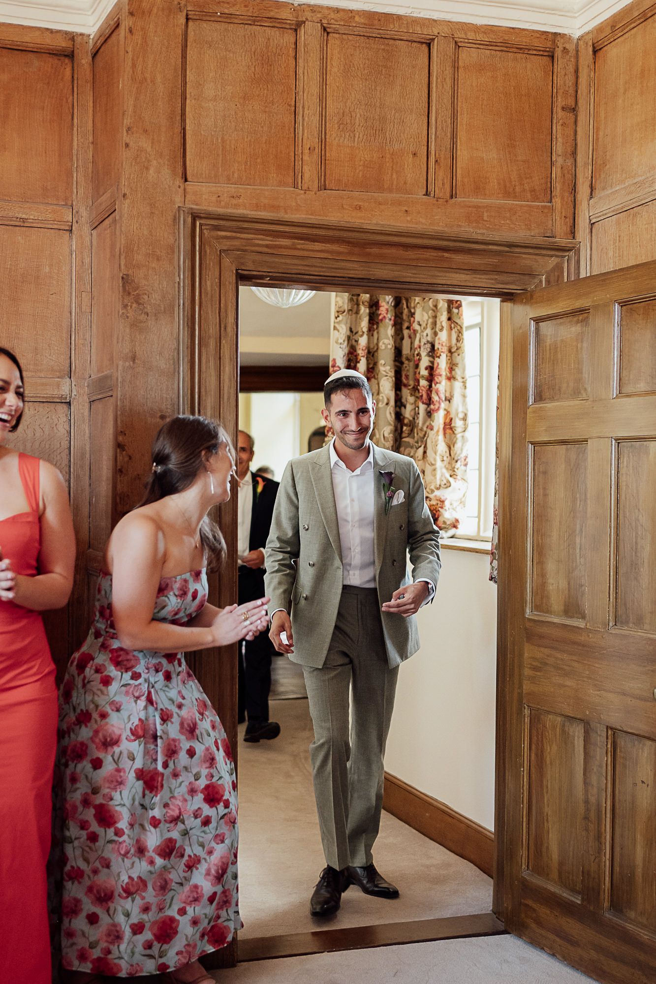 Smiling groom in sage suit enters wood-paneled room greeted by wedding guests in colorful dresses.