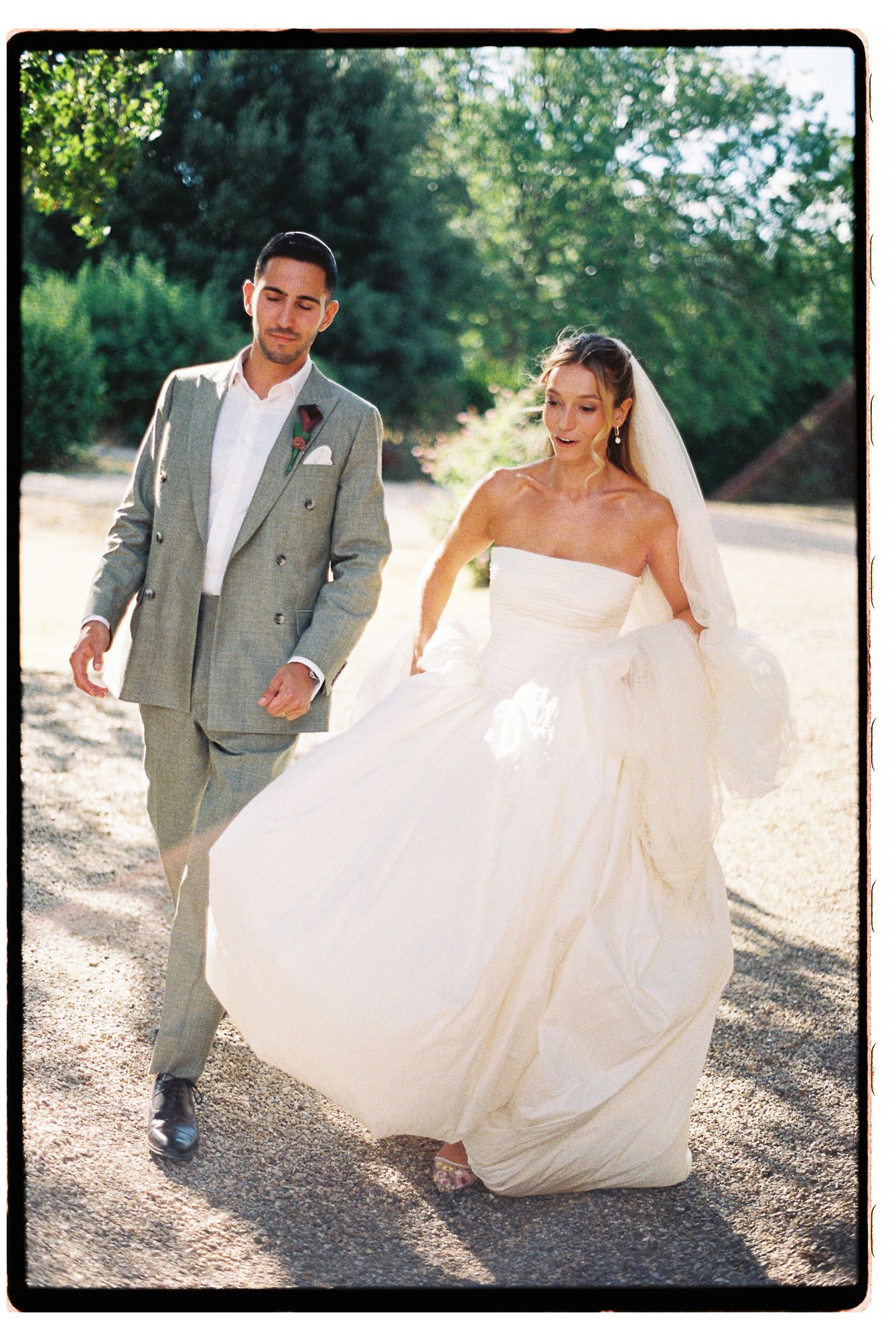A bride in a flowing white strapless gown and veil walks with a groom in a gray plaid suit on a sunny outdoor path.