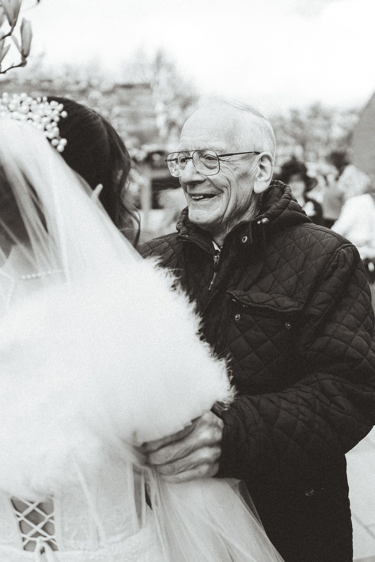 Black and white photo capturing a joyful moment between a bride in her veil and a man in glasses at a wedding celebration.