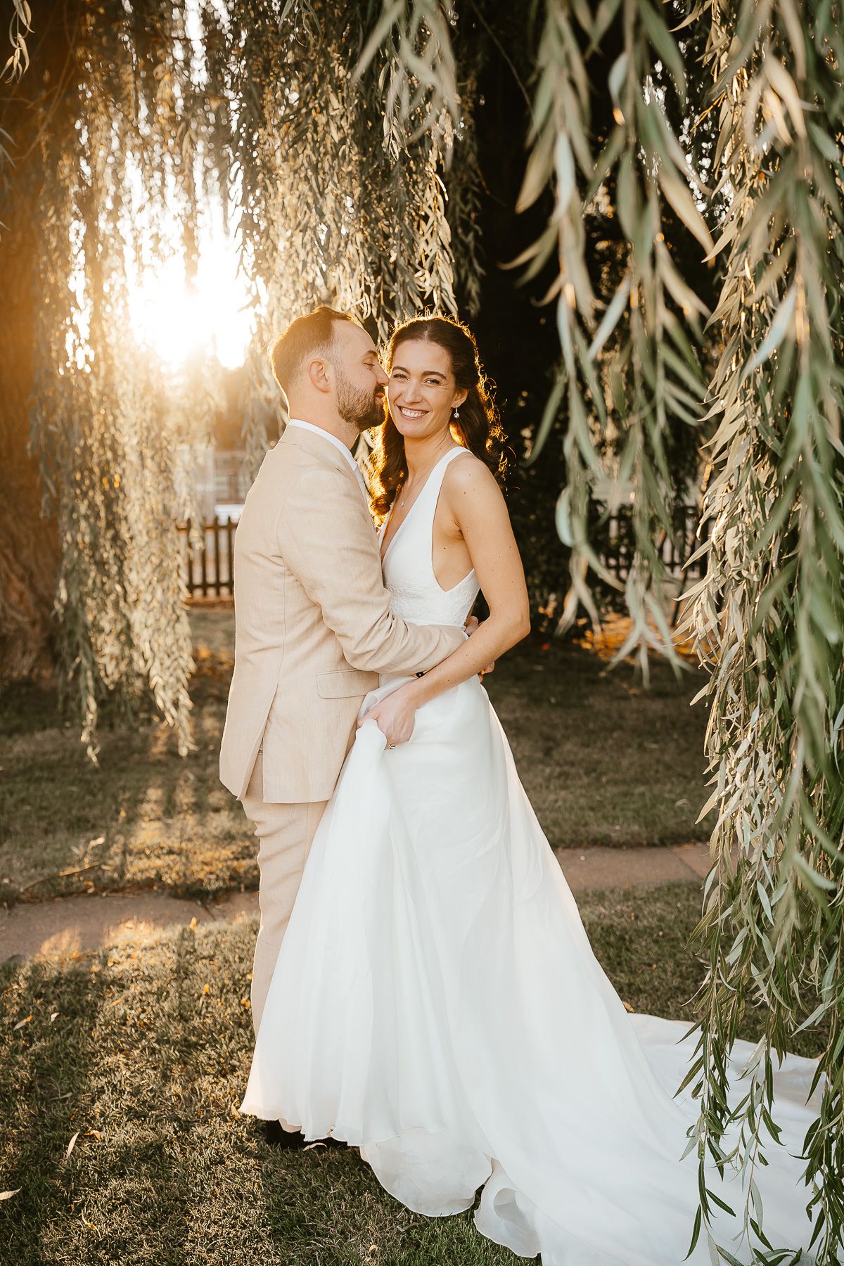 A romantic sunset wedding photo under a weeping willow tree with golden light filtering through hanging branches.