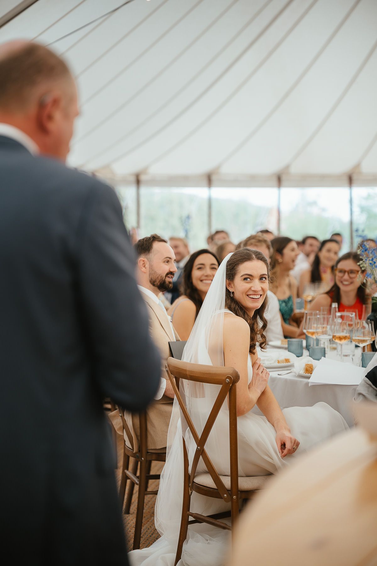 Wedding guests and a bride in a white dress sit at tables under an elegant white tent during a reception celebration.