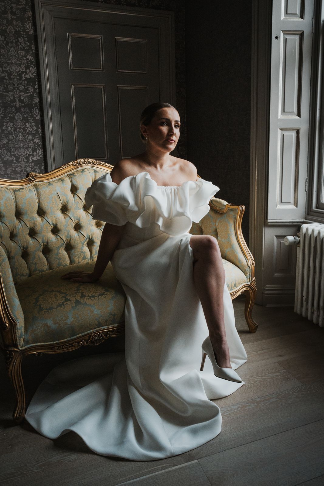 Elegant off-shoulder white wedding gown with ruffles photographed in moody lighting on vintage tufted sofa.