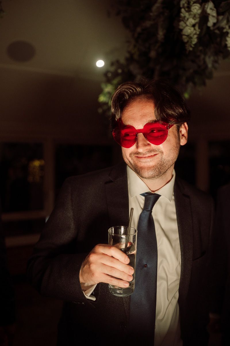 A person in a suit and red sunglasses holds a champagne glass in a dimly lit setting.