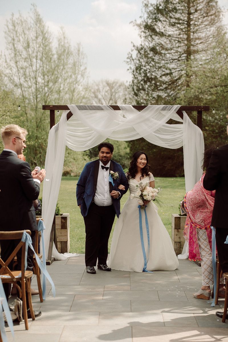 Wedding ceremony setup outdoors with white draped fabric arch and wooden chairs decorated with light blue ribbons.
