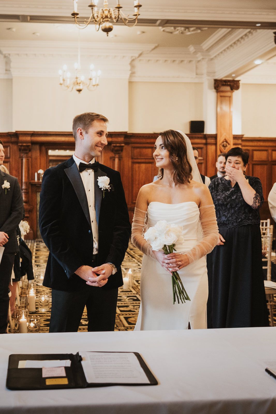 Wedding couple signs marriage documents at elegant ceremony with wood paneling and chandeliers.