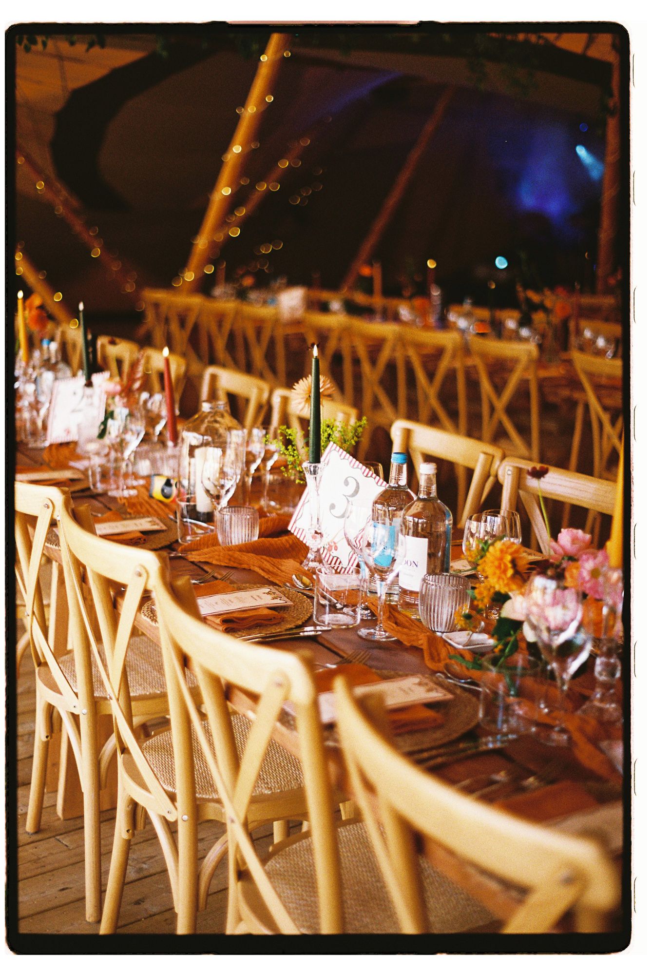 Elegant tipi wedding reception with long wooden tables, cross-back chairs, and colorful floral centerpieces.