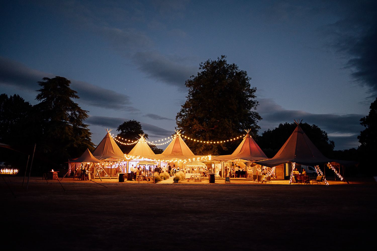 Illuminated wedding marquee tents with festoon lights glowing warmly against a twilight sky surrounded by trees.